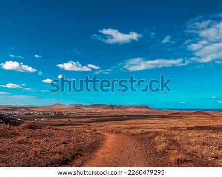 Trail towards town in arid land. Skyline of mountains in the background. A little town with white houses before the mountains. Desertic vegetation.