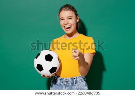 Young woman fan wear basic yellow t-shirt cheer up support football sport team hold in hand soccer ball watch tv live stream point index finger camera you encourage isolated on dark green background Royalty-Free Stock Photo #2260486905