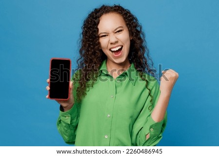 Young fun woman of African American ethnicity 20s she wear green shirt hold in hand use mobile cell phone with blank screen workspace area do winner gesture isolated on plain blue background studio