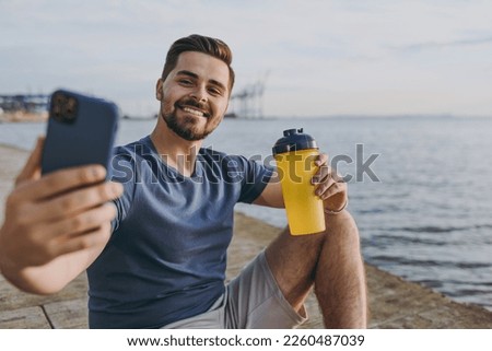 Young fun sporty strong sportsman man in sports clothes shorts train do selfie shot photo on mobile cell phone drink water at sunrise sun over sea beach outdoor on pier seaside in summer day morning