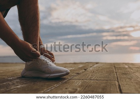 Close up cropped photo shot of sportsman man wearing sports clothes warm up training sit laces up sneakers shoes at sunrise sun dawn sky over sea beach on pier outdoor seaside in summer day morning