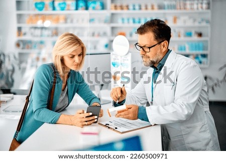 Mid adult woman using cell phone while consulting with her pharmacist in choosing medicine in a pharmacy. Royalty-Free Stock Photo #2260500971