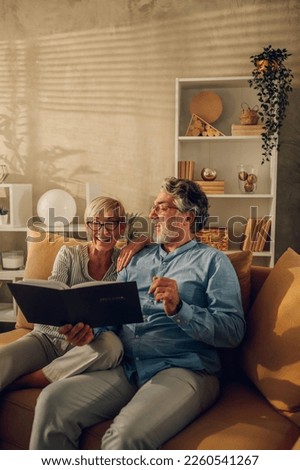 Smiling senior couple looking into their photo album while sitting on a couch at home. Mature spouses talking of marriage history and raising their children trough the years. Copy space.