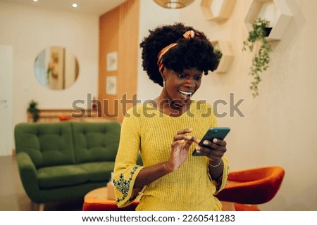 Smiling black woman using smartphone at home. African american girl typing a message on a mobile phone or replaying an email. Connection, mobile apps and lifestyle concept. Copy space. Royalty-Free Stock Photo #2260541283