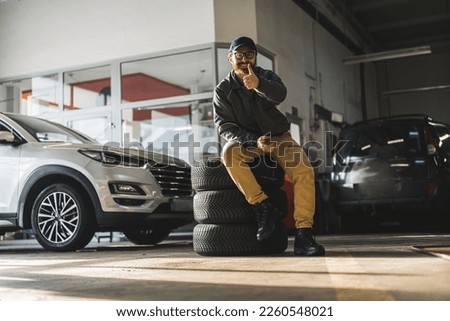 a mechanic with glasses sitting on wheels and showing thumb-up in a car repair shop, full shot. High quality photo Royalty-Free Stock Photo #2260548021