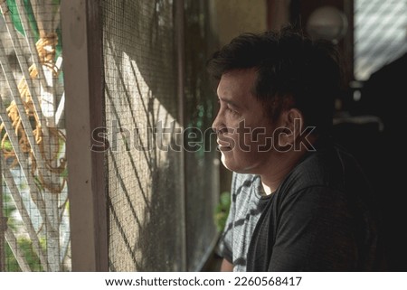 A saddened middle aged man peers through the screen window, longing for a loved one. Indoor dramatic scene. Royalty-Free Stock Photo #2260568417
