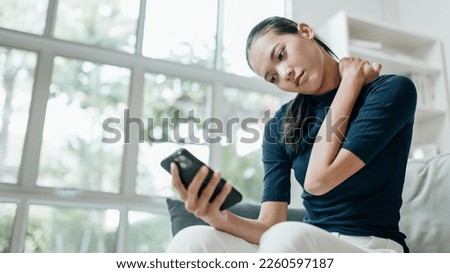 Portrait of Unhappy asian woman suffering from pain in the neck while sitting on the sofa and using mobile phone in the living room. Lifestyle, health care and medical concepts. Royalty-Free Stock Photo #2260597187