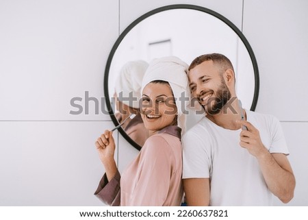 Cheerful caucasian young couple at bathroom brushing teeth toothy smiling cuddling. Newlyweds enjoying honeymoon, at hotel room. Pretty hispanic girl with towel on head having fun with boyfriend.