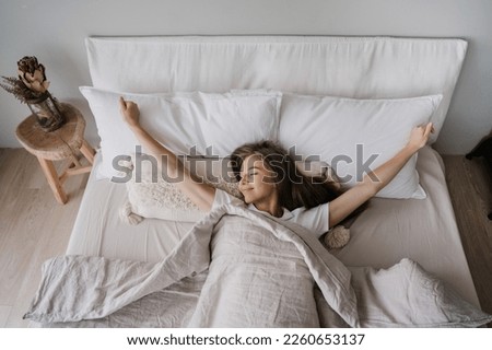 Young long haired Italian girl laying on bed eyes closed awakening at Sunday morning smiles satisfied by healthy sleep. Top view on young woman spreading hands, stretching. Cozy home, happy people. Royalty-Free Stock Photo #2260653137