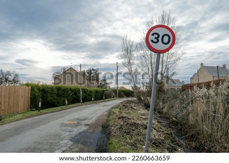 Bent over 30 Mph speed limit sign seen approaching a small, rural village. Know for speeding cars, the road surface is heavily damaged due to tractors.