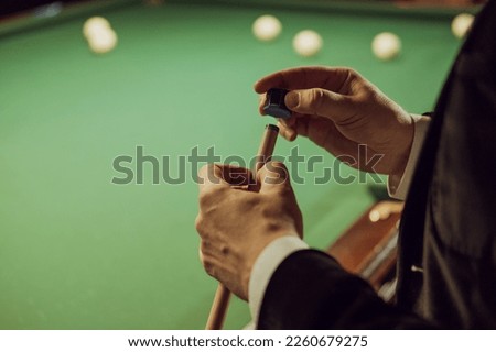 Close up of man in a jacket rubs the end of a cue with chalk before the game. Background of green pool table with billiard balls Royalty-Free Stock Photo #2260679275