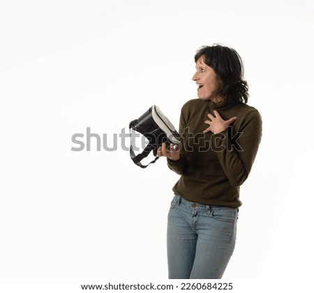 Middle aged Caucasian girl making surprised gesture looking towards copy space with virtual reality goggles in hand. Dressed in casual clothes on white background.