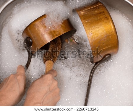 A pair of lady's hands washing a copper pan with a wooden brush whilst another copper pan soaks in a frothy sink of warm water Royalty-Free Stock Photo #2260713245