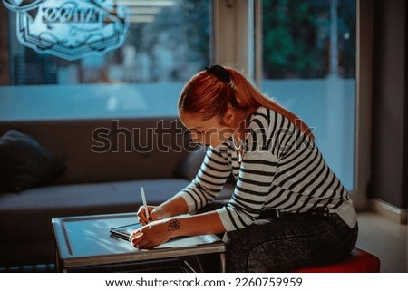 Tattoo artist with red hair drawing on her tablet while sitting at the studio