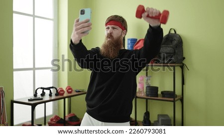 Young redhead man using dumbbells training and making selfie by smartphone at sport center