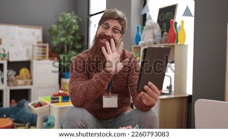 Young redhead man preschool teacher having video call sitting on chair at kindergarten
