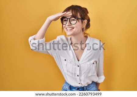 Young beautiful woman wearing casual shirt over yellow background very happy and smiling looking far away with hand over head. searching concept.  Royalty-Free Stock Photo #2260793499