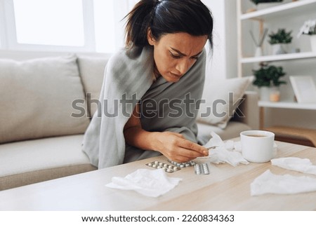 A woman with a cold with pills is treated at home chooses which drugs to take and self-medicates, checks the expiration date while sitting on the couch at home Royalty-Free Stock Photo #2260834363