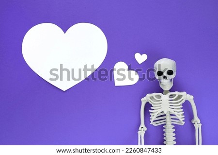 Skeleton on violet background with white blank paper cut hearts, one big heart with copy space. Anatomical plastic model human skeleton with dreaming text balloon. Valentine's day love concept.