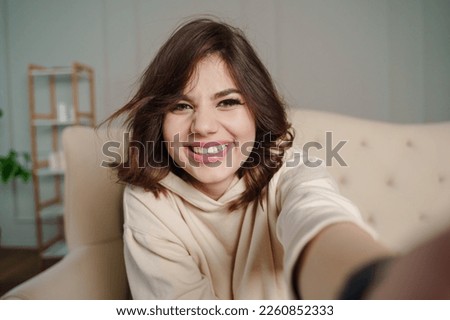 Pretty young female with big smile sitting at living room. She having fun taking selfie.