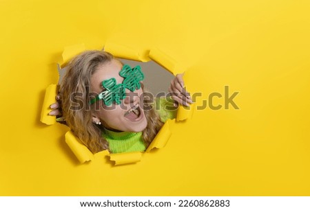 Surprised woman wearing green clover-shaped glasses looking, peeping through the bright yellow paper hole. Showing hand to side. Copy space for text. St Patrick day. Wow face  

