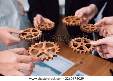 Closeup top view businesspeople hand holding gear and join together over meeting table with financial report papers. Cohesive group of office workers holding cog wheel as synergy harmony concept.