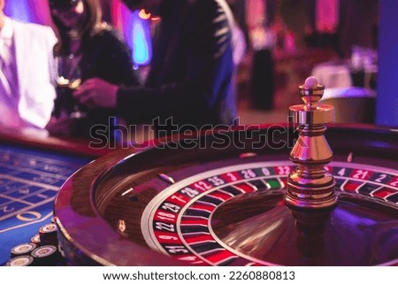 Vibrant casino table with roulette in motion, with casino chips, tokens, the hand of croupier, dollar bill money and a group of gambling rich wealthy people playing bet in the background
 Royalty-Free Stock Photo #2260880813
