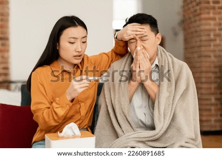 Loving young asian wife taking care of her sick middle aged husband, checking fever with thermometer, touching his forehead, sitting on sofa at home. Korean man having flu, sneezing nose Royalty-Free Stock Photo #2260891685