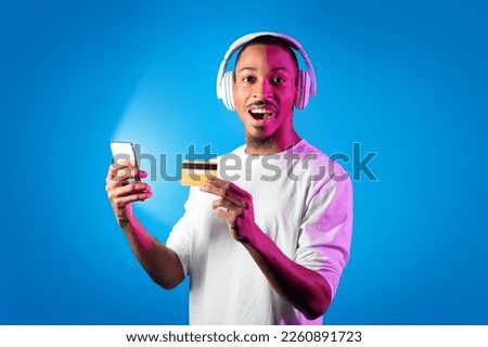 Newest musical app concept. Excited handsome young black guy in white longsleeve using modern smartphone with shining screen, wireless headphones and credit card over studio background in neon light