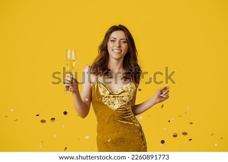 Posh party. Glamour young lady in festive evening dress standing with glass of champagne under falling confetti and smiling at camera, yellow studio background Royalty-Free Stock Photo #2260891773