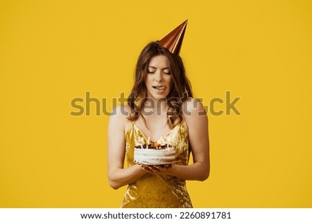 Pretty woman in dress licking her lips from temptation, looking at delicious birthday cake with yearning to bite it, standing with dessert against yellow background