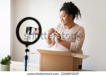 Beauty blogger cute young hispanic curly woman plus size unpacking parcel at home, recording video for her blog, using blogger set phone and tripod with led lamp, reading beauty product label