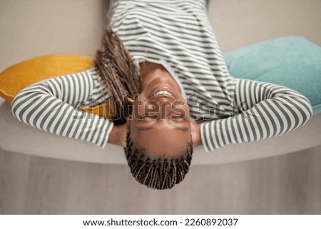 Top view of happy joyful pretty young black woman in casual outfit chilling on couch at home, reclining on sofa with closed eyes and arms behind her head, dreaming about wealthy future, closeup