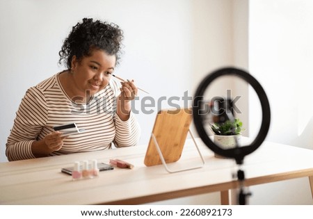 Beauty blog, blogging concept. Cheerful pretty curly young mixed race plump woman in casual outfit sitting at desk in front of mirror at home, recording video while applying makeup, using blogger set
