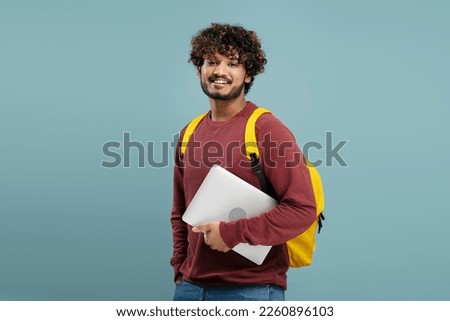 Attractive smiling Indian student with yellow backpack holding laptop,  looking at camera isolated on blue background. Education concept 