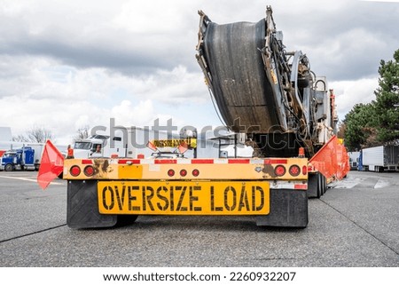 Industrial powerful big rig classic semi truck transporting oversized equipment on step down semi trailer with oversize load sign on the back standing for the rest on the truck stop parking lot Royalty-Free Stock Photo #2260932207
