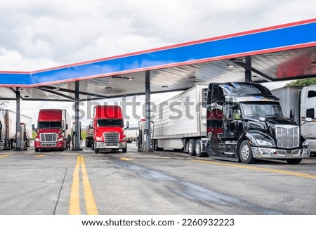 Industrial standard big rigs semi trucks with semi trailers standing on the fuel station parking lot filling the semi trucks tanks with diesel fuel to have ability continue commercial flight Royalty-Free Stock Photo #2260932223