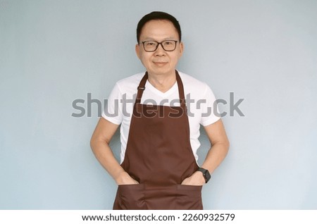 Mature confident successful small business owner Asian man wearing glasses, brown apron and looking at camera isolated green pastel color background. Start up small business entrepreneur SME concept.