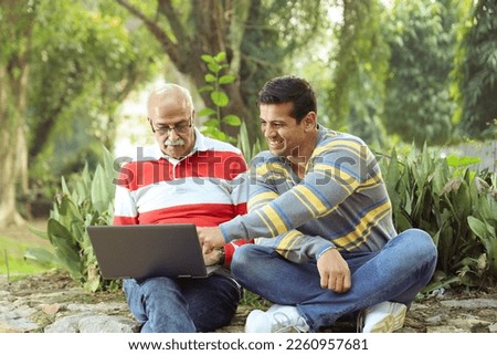 Happy Indian old man sitting in a lush green atmosphere in a park holding a laptop with his grandson, making the best use of technology and internet and planning vacations.