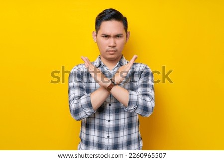 Serious young handsome Asian man wearing a plaid shirt crossing his hands makes stop gesture, demonstrates rejection isolated on yellow background. People lifestyle concept