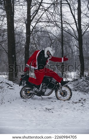 A motorcyclist in a Santa Claus suit funny jumping. Winter forest with falling snow. Motorcycle in the background. The concept of New Year's holidays and sales. Vertical photo.