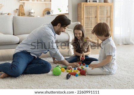 Happy daddy engaged in game with cheerful little son and daughter kids, playing toy blocks on soft carpeted floor, arranging cubes from heap, talking, smiling, laughing, enjoying leisure, playtime
