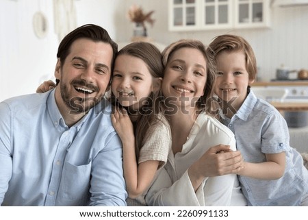 Happy attractive young couple of parents and cute cheerful little kids hugging, sitting close in row, looking at camera, smiling, laughing, enjoying family shooting, closeness, having fun