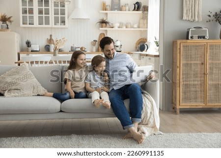 Cheerful millennial dad entertaining two little kids at home, resting on couch, using laptop computer, making video call, watching funny movie online, enjoying Internet communication