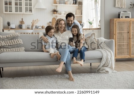 Cheerful mom, dad and two little kids using mobile phone at home together, hugging on sofa, playing online virtual game, taking self video for blog, social media, making family call Royalty-Free Stock Photo #2260991149
