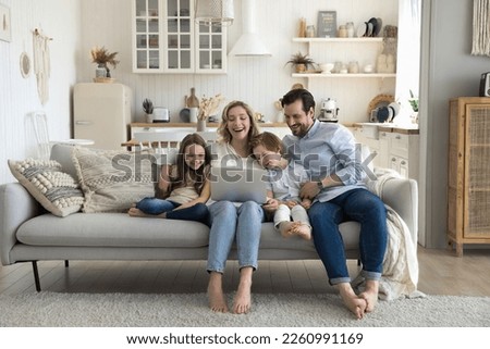 Two cheerful little kids, happy mom and dad enjoying leisure at home together, using laptop on sofa, talking on video call, laughing, watching movie, interactive online TV channel