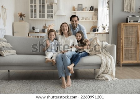 Joyful family couple and little son and daughter kids sitting on sofa, resting on comfortable couch at cozy home, hugging with love, joy, looking at camera, smiling. Full length wide shot