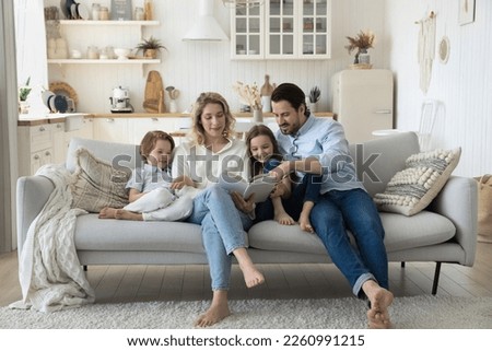 Positive engaged couple of parents teaching two cute kids to read book, watching pictures, telling funny fairytale, relaxing on home couch, hugging children, enjoying literature hobby Royalty-Free Stock Photo #2260991215