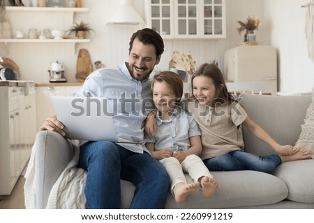 Happy father and two joyful little kids using media app on laptop for Internet communication, watching cartoon funny movie, online TV family channel, resting on sofa at home