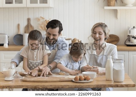 Cheerful happy couple of parents and two cute little kids in aprons teaching to bake, rolling, shaping dough. Children helping mom and dad to prepare dessert, pie, pastry food, Royalty-Free Stock Photo #2260991263